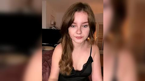 Media: Video of a young woman with fair skin and long brown hair, wearing a black camisole, sitting on a bed with a blurred background of a bedroom.