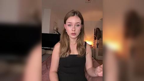 Media: Video of a young woman with fair skin and light brown hair, wearing a black tank top, sitting on a pink rug in a dimly-lit room with a warm glow.