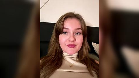 Media: Video of a Caucasian woman with long brown hair, light skin, and light pink lipstick, wearing a beige turtleneck sweater. Background includes a black leather couch and beige walls.