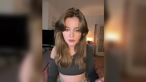 Media: A blurry video of a young woman with wavy brown hair, wearing a black crop top, in a dimly-lit bedroom with a TV, lamp, and closed blinds in the background.