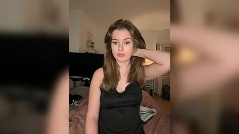 Media: A video of a young woman with long brown hair, wearing a black tank top, posing in a bedroom with messy furniture and a blurred figure in the background.