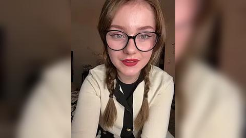 Media: Video of a fair-skinned woman with braided hair, wearing glasses, a beige cardigan, and black top, sitting indoors. Background is blurred.