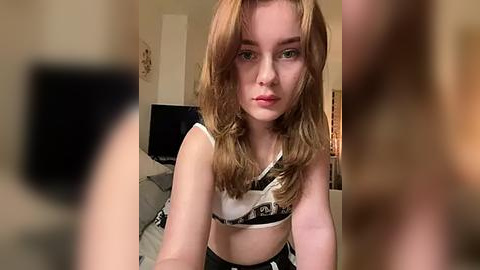 Media: Video of a fair-skinned, young woman with long, wavy, light brown hair, wearing a black and white bikini top, sitting on a bed with gray sheets in a dimly lit room.