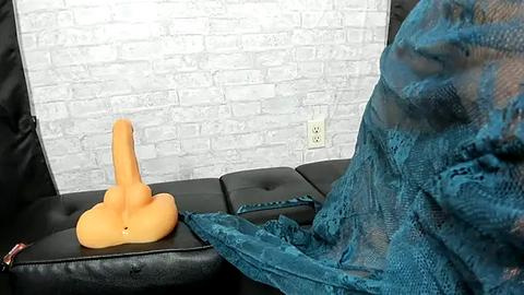 Media: Video of a plush, orange, penis-shaped toy on a black leather couch, draped in a teal lace blanket against a white brick wall background.