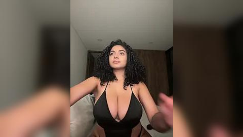 Media: A video of a curvy woman with dark curly hair, wearing a plunging black swimsuit, taking a selfie in a dimly lit bedroom.