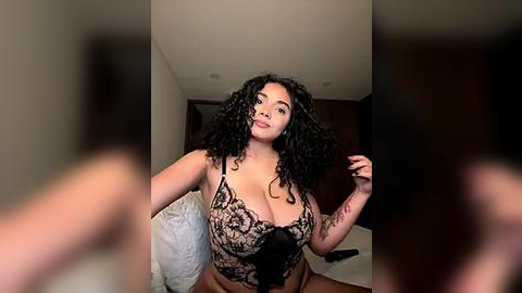 Media: Video of a curvy Black woman with long curly hair, wearing a black lace bodysuit, standing in a bedroom with white bedding and dark wooden walls.