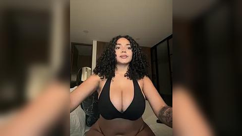 Media: Video of a plus-sized woman with curly black hair, large breasts, and brown skin, wearing a black halter top, sitting on a bed in a modern, dimly lit bedroom.