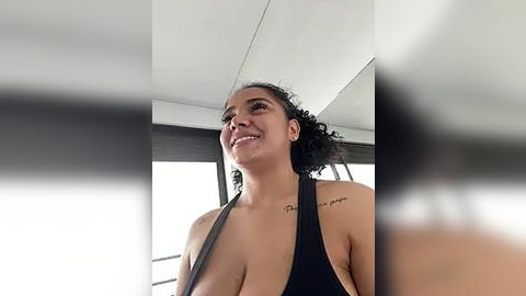 Media: Video of a smiling, curly-haired woman with medium skin tone, wearing a black halter top, with a tattoo on her left shoulder, standing indoors next to a window.