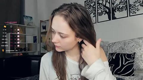 Media: Video of a young woman with light skin and long brown hair, wearing a white long-sleeve shirt, adjusting her earbud in a modern, minimalist living room with white walls and black tree art.