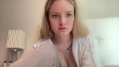 Media: Video of a fair-skinned, blonde woman with a septum piercing, wearing a white shirt, sitting on a bed. Background features a white lampshade and blurred white walls.