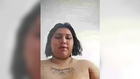 Media: A grainy video of a naked woman with shoulder-length black hair, medium skin tone, and a prominent tattoo across her chest. The background is blurred.