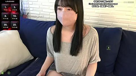 Media: A video shows a young Asian woman with long black hair, wearing a grey top, sitting on a dark blue couch in a living room with white brick walls.