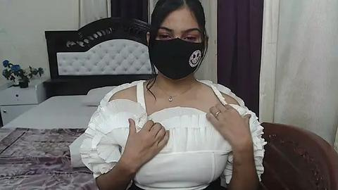 Media: Video of a woman with medium skin tone, wearing a black mask, white ruffled blouse, and black top, sitting on a bed with white and purple linens.