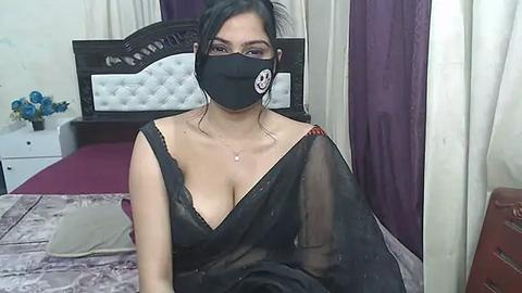 Media: Video of an Asian woman with medium skin tone, black mask, black dress, sitting on a bed with purple curtains, white headboard, and floral decor.