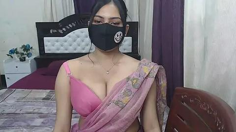 Media: Video of a young South Asian woman in a pink bra, black face mask, and pink saree, seated on a bed with white headboard and purple curtains, in a simple bedroom.