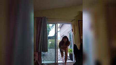 Media: Video of a woman with long hair, wearing a black top, standing in a doorway, with a patio and garden visible through a sliding glass door.