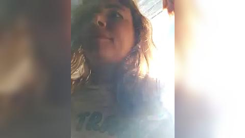 Media: A video of a young woman with long, wavy hair, wearing a gray t-shirt, captured from a low angle with bright light causing a halo effect around her head.