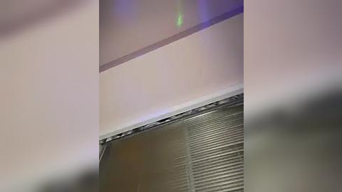 Media: Video of a ceiling with a white border, beige wall, and a metal vent cover with horizontal slats partially visible. The image has a slight blur, making it difficult to discern fine details.