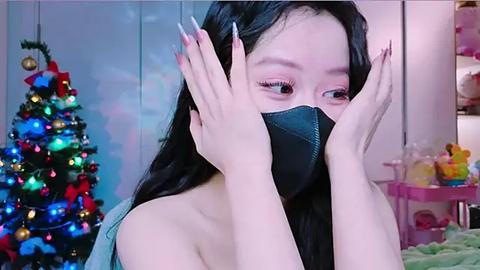 Media: Video of an East Asian woman with long black hair, wearing a black mask and light blue top, playfully covering her face, in a festive room with a decorated Christmas tree, toys, and soft pink background.