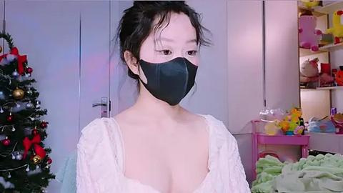 Media: Video of an East Asian woman with fair skin, black hair tied up, wearing a black mask and white lace top, standing indoors with a Christmas tree, toys, and shelves in the background.