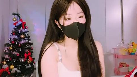 Media: Video of an Asian woman with long black hair, wearing a black face mask, white tank top, and red earrings. Background includes a decorated Christmas tree and a shelf with toys.