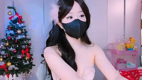 Media: Video of an Asian woman with long black hair, wearing a black mask, light-colored dress, and white headband, standing in front of a decorated Christmas tree and pink storage shelves.