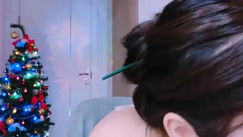 Media: Video of a woman with dark hair styled in a bun, wearing green hairpins. She stands in front of a light blue door with a Christmas tree ornament on it, featuring colorful baubles and a red bow.