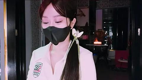 Media: Video of an Asian woman in a black face mask, white shirt, and long dark hair with a white flower, standing indoors with dim lighting, blurred background of a cozy room.