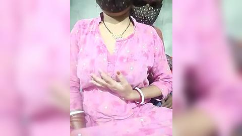 Media: Video of a woman in a pink floral dress, seated, wearing a black face mask and a silver necklace, with a blurred background.