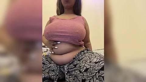 Media: A video of a plus-sized woman with medium brown skin, wearing a striped pink crop top and patterned black pants, sitting on a bed. Her large breasts are partially lifted by her hand. The background is blurred.