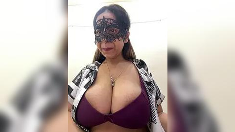 Media: Video of a light-skinned woman with long brown hair, wearing a black lace masquerade mask, revealing a burgundy bra highlighting her large breasts, and a black-and-white checkered shirt. The background is a plain white wall, and there is a blurred out person in the foreground.