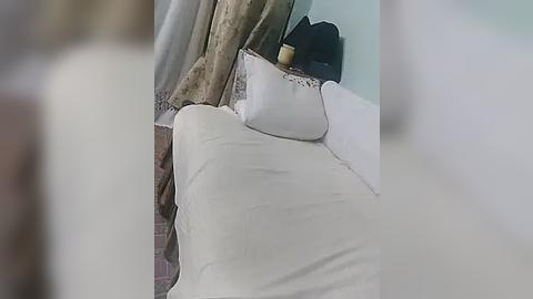 Media: Video of a neatly made bed with white sheets and a beige pillow, adorned with a small, black, round object. The background shows a partially visible wall with beige curtains. The scene is softly lit, suggesting a serene, minimalist bedroom.