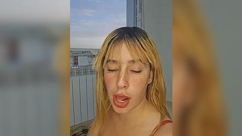 Media: Video of a young woman with long, blonde hair, light skin, and a red top, smiling and licking her lips. Background shows a blurred cityscape.