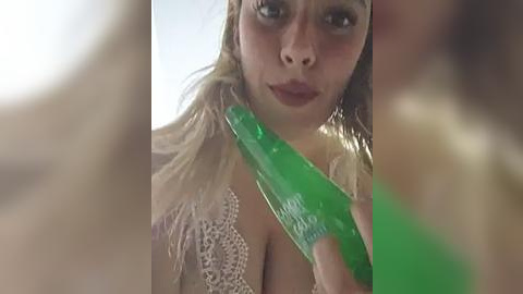 Media: Video of a woman with wet, blonde hair, wearing a lacy white top, holding a green toothbrush against her cheek, mouth slightly open, blurry background.