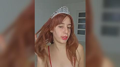 Media: A video of a young woman with fair skin and reddish-brown hair, wearing a silver tiara and a red spaghetti-strap top, adjusting her hair in a softly lit room.