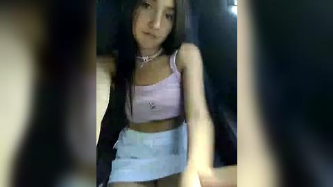 Media: A video of a young woman with long black hair, wearing a light purple crop top and high-waisted denim shorts, seated indoors, blurred foreground.