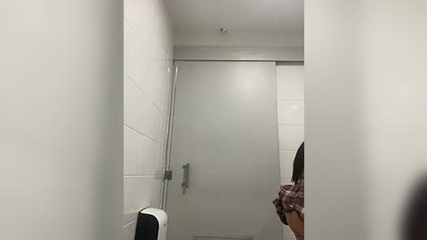 Media: Video of a small, modern bathroom with white tiled walls, a shower with a glass door, a black and white hand dryer on the left, and a partially visible person in a plaid shirt.