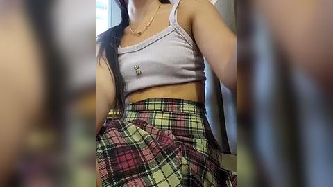 Media: Video of a young woman with long black hair, wearing a white crop top and a high-waisted plaid skirt, posing indoors.