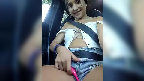 Media: Video of a young woman with fair skin and brown hair in a car, wearing a white off-shoulder crop top and denim shorts, driving while using a pink vibrator.