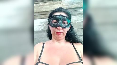 Media: Video of a woman with long black hair, wearing a green lace masquerade mask and a black strappy top, standing in front of a rustic wooden wall.