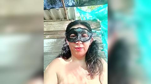 Media: Video of a topless woman with long dark hair, wearing a black masquerade mask and red lipstick, standing in a rustic, outdoor setting with wooden walls and a blue tarpaulin.