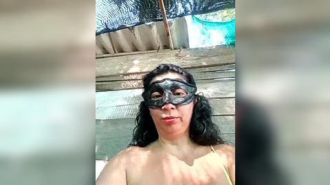 Media: A video of a woman with long black hair, wearing a black masquerade mask, standing in front of a rustic wooden structure with a blue pool in the background.