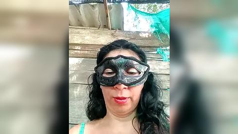 Media: A video of a woman with long black hair and light skin wearing a black, lace masquerade mask with teal accents, standing against a rustic wooden fence with green netting in the background.