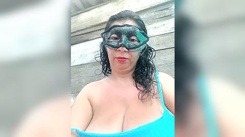 Media: Video of a curvy woman with long black hair, wearing a blue tank top and a green masquerade mask, standing in front of a wooden wall.