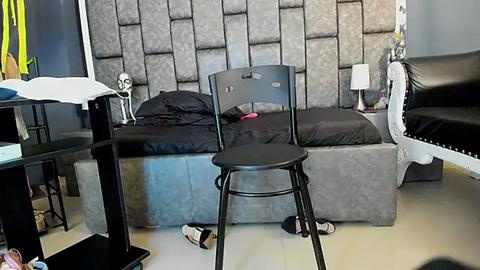 Media: Video of a contemporary bedroom with a gray, tufted headboard bed, a black stool, a black chair, a white lamp, and a black-and-white striped bedspread.