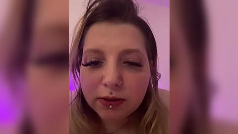 Media: Video of a young woman with light skin, brown hair, and multiple facial piercings, including a septum ring and lip rings. She has a neutral expression and is indoors with blurred purple lighting in the background.