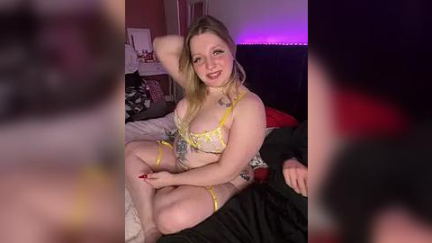 Media: A video of a blonde, fair-skinned woman in yellow lingerie with a tattooed arm, seated on a bed with a purple light, surrounded by blurred figures.