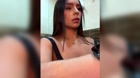 Media: A close-up video of a young woman with long, straight brown hair, wearing a black tank top, standing in a bathroom with beige tiles and a dark countertop.