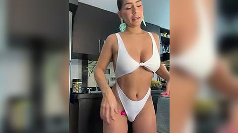 Media: Video of a fit, tan-skinned woman with short dark hair, wearing a white high-cut bikini, standing in a modern kitchen.