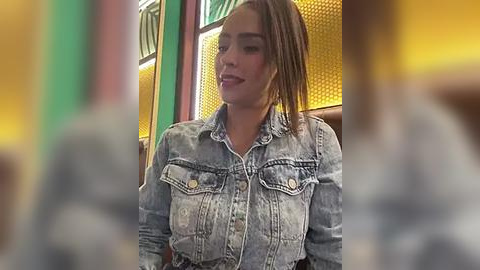 Media: Video of a young woman with light skin and shoulder-length brown hair in a denim shirt, seated indoors near a window with yellow-tinted glass.